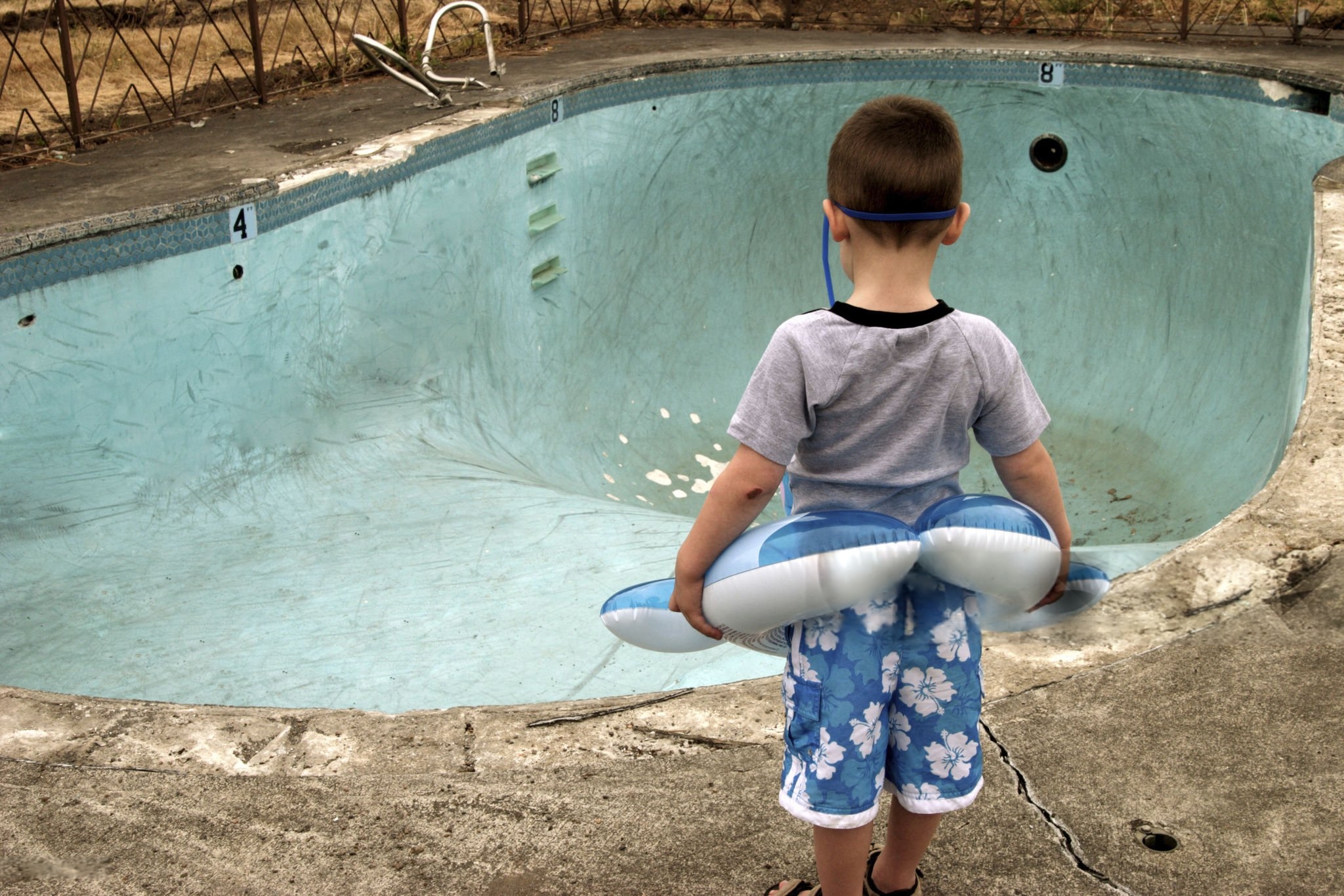 Child looking at an empty, damaged swimming pool with visible cracks and peeling surface